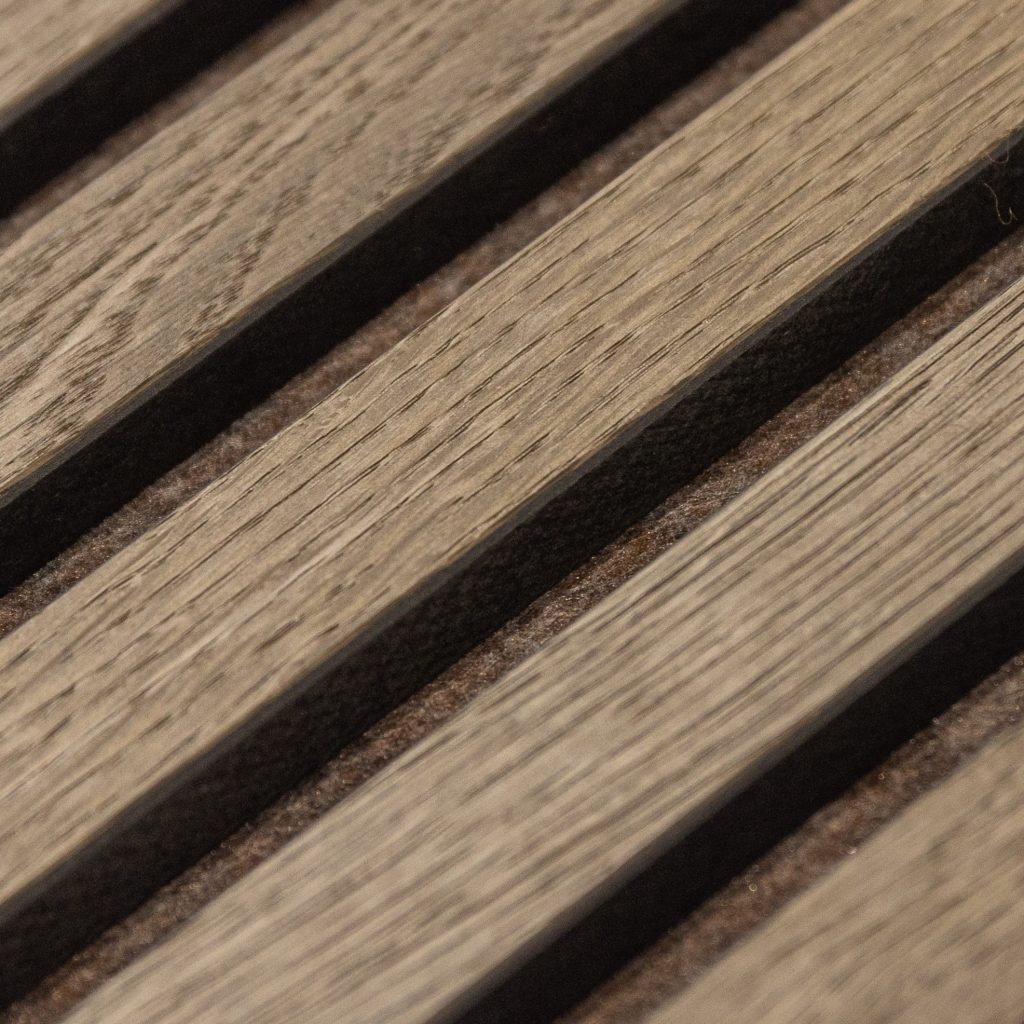 Nært billede af WoodUpp akustiklameller i gråbrun egefinér, der fremhæver træstruktur og diagonalt placerede smalle lameller med mørk filtbaggrund i sporene. Detaljer viser fine årer, svagt børstet mat overflade, naturlige nuancer og præcise mellemrum — WoodUpps dekorative, lydabsorberende vægpaneler til indendørs brug.