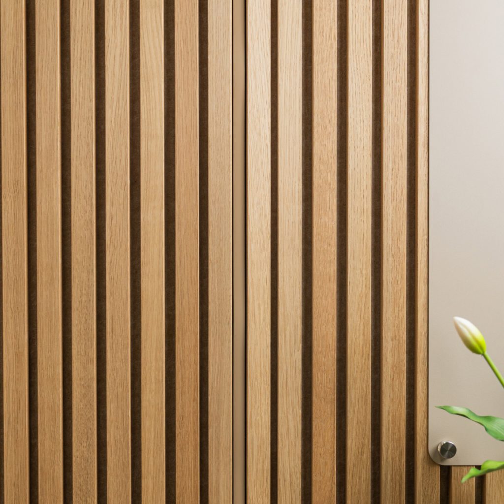 WoodUpp lyst egetræslamelpanel med smalle, lodrette lameller og mørke fuger, monteret som vægbeklædning. Billedet viser tætstående trælameller med glat overflade, en del af en lysegrå metalplade med rund fastgørelse og en hvid tulipanknop i nederste højre hjørne. Naturlig åretegning og varm, mat finish.