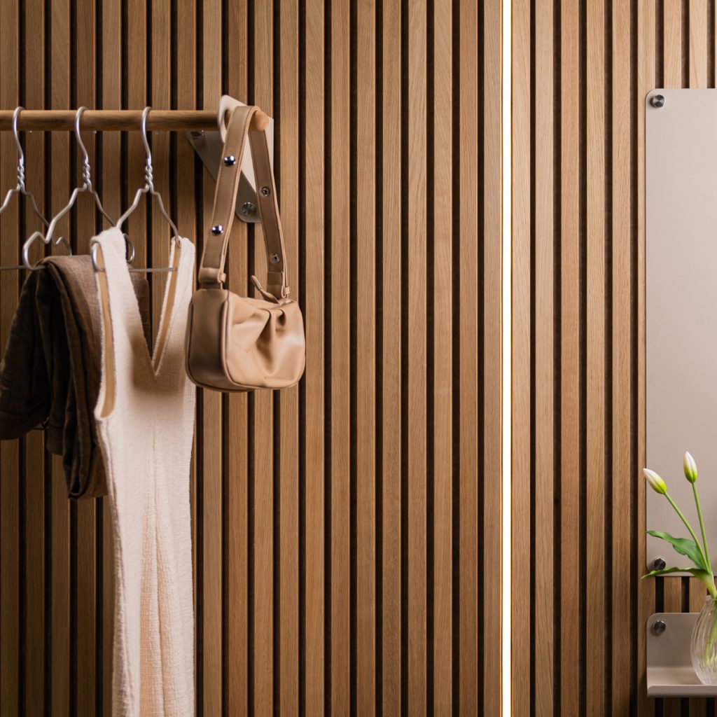 WoodUpp lamelvæg i lyst træ med tætte, lodrette lameller; WoodUpp knager og metalbeslag bærer en træstang med bøjler, tøj og en lille taske; til højre ses et WoodUpp spejl med synlige afstandsbeslag og en lille hylde med vase i varmt, blødt lys.