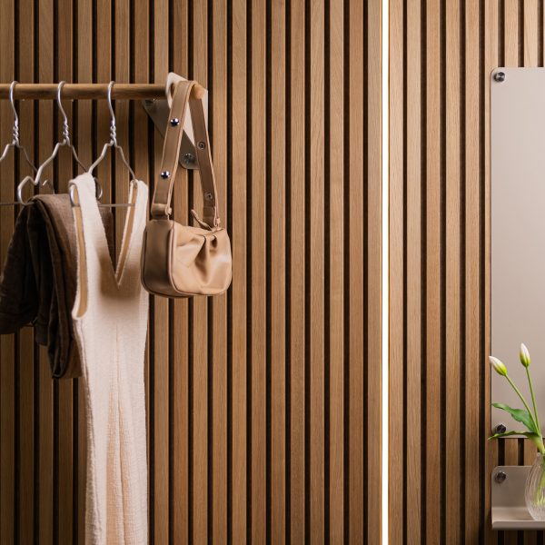 WoodUpp lamelvæg i lyst træ med tætte, lodrette lameller; WoodUpp knager og metalbeslag bærer en træstang med bøjler, tøj og en lille taske; til højre ses et WoodUpp spejl med synlige afstandsbeslag og en lille hylde med vase i varmt, blødt lys.