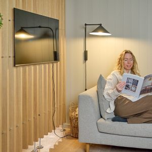 Kvinde sidder på en grå sofa og læser et magasin under en væglampe, mens en fladskærm er monteret på WoodUpps lodrette trælameller/vægpaneler i lys eg, der danner en dekorativ og akustisk bagvæg, med synlig strømledning, kurv med tæpper og metalføtter ved panelerne.