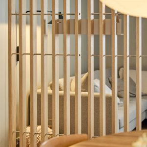 Billede af WoodUpp skillevæg bestående af smalle, lodrette egetræslamel-paneler med synlige WoodUpp metalbeslag og tværstænger i en varm, naturlig egetræsfinish, der deler et lyst opholdsområde. Bagved anes en beige sofa med puder og væglamper, set gennem jævnt placerede trælameller, hvilket skaber et moderne, indbydende rum.
