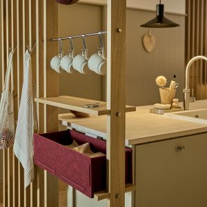 WoodUpp-modul i egetræsramme viser et køkkenafskærmende reolsystem med lodrette lameller, hylde og rustfri stang med fem hvide kaffekopper, en dyb bordeaux filt-skuffe og små træknopper. Scenen viser WoodUpps varme, funktionelle opbevaringsløsning i naturlige materialer. Lys egetræsbordplade, køkkenø med synligt vaskarmatur samt netpose og stribet viskestykke hængende ved siden.