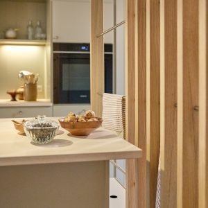 WoodUpp: Nærbillede af et lyst køkken med WoodUpp lamelvæg i egetræ, integrerede metal håndklædeholdere og en lys træbordplade ved køkkenøen. På bordet står træskåle med rodfrugter og en gennemsigtig glaskrukke; i baggrunden ses ovn og oplyste skabe. Varmt lys kaster blødt skær over overfladerne.