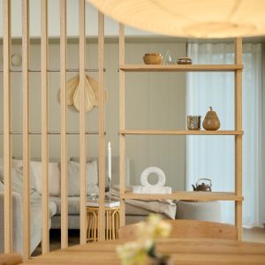WoodUpp rumdeler i lyst egetræ med vertikale lameller og indbyggede hylder, vist som åben reol med små dekorative genstande: træskål, glasfigur, keramisk pære, marmorring og opstilt lys — i en lys, neutral stue med sofa og bløde gardiner og naturlig varme.