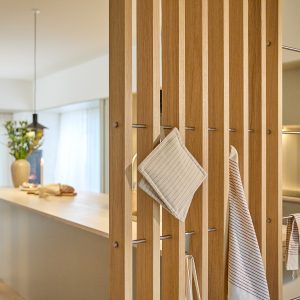 WoodUpp trælamelvæg i lyst egetræ med vertikale lameller som rumsdeler, udstyret med slanke polerede metalstænger/kroge fra WoodUpp til praktisk ophæng. På dem hænger grydelapper og stribede viskestykker; bagved ses et lyst køkkenø og bløde gardiner i en varm, minimalistisk indretning.