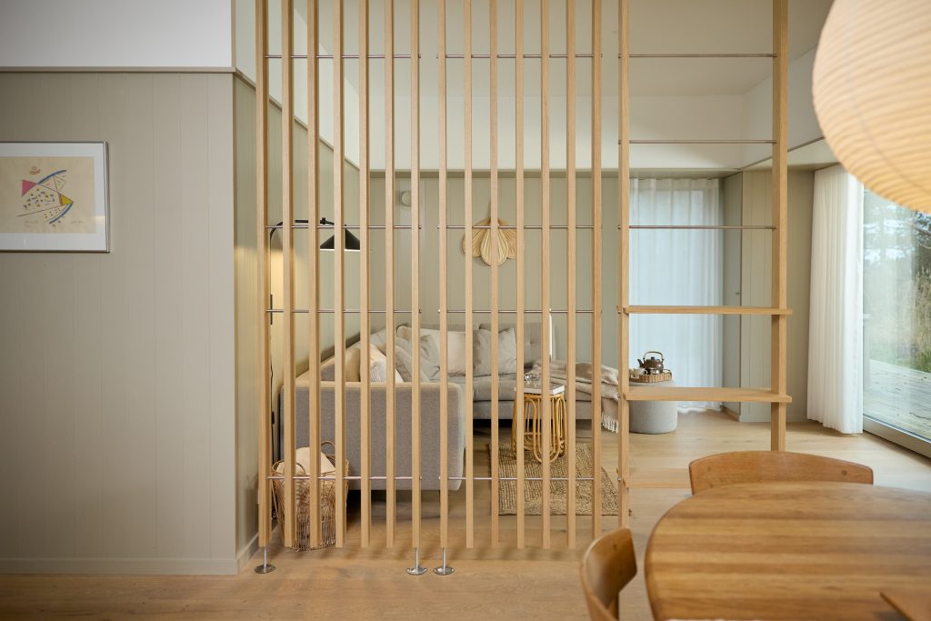 WoodUpp rumdeler bestående af lyse, lodrette egetræslameller med metalstangforbindelser, der deler spise- og opholdsområde. Til højre ses en WoodUpp åben hylde/reol i samme træ. Bag lamellerne står grå sofa, kurv, rattan sidebord, sort væglampe og store vinduespartier med hvide gardiner.
