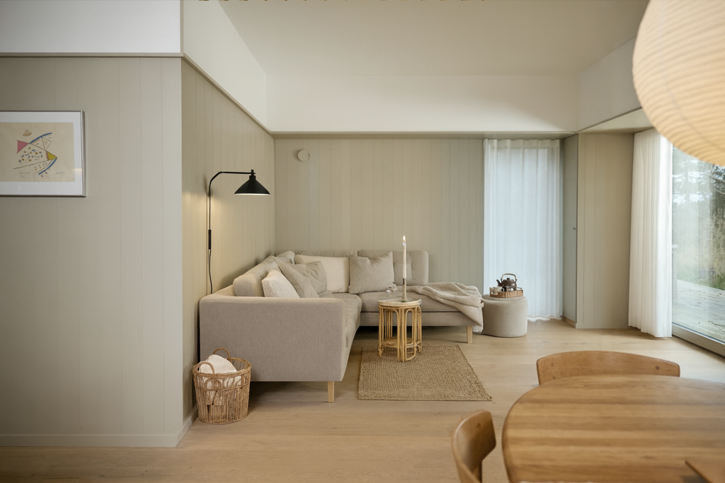 Billedet viser en hyggelig stue indrettet med WoodUpp vægpaneler. Væggene er dækket af malede, lodrette træpaneler i en lys beige farve, der skaber en rolig atmosfære. Der er en beige hjørnesofa, et lille bambusbord og et spisebord i træ i forgrunden. Rummet er lyst og nordisk.