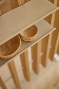 Detaljeret foto af en WoodUpp-rumsdeler med lodrette, lyse trælameller og en smal beige WoodUpp-hylde, hvor to små, runde drejede træskåle ligger ved siden af hinanden. Billedet viser kornet i træet, hyldens matte overflade og blød, varm belysning i naturlige nuancer og minimalistisk design.