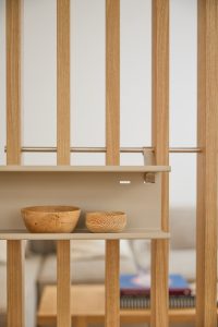 WoodUpp rumdeler i lyst egetræ med lodrette lameller og en aftagelig, beige metalhylde monteret midt på. På hylden står to WoodUpp hånddrejede træskåle i forskellig størrelse med tydelige årer og naturlig overflade. Enkel skandinavisk æstetik, naturtoner og minimalistisk opbevaring, velegnet til entré eller som rumafgrænsning i stuen.