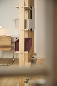 WoodUpps lyse egetræ-rumdeler med vertikale lameller og synlige skruer, hvor to WoodUpp filtkasser — en lys grå og en dyb vinrød — sidder som enkle opbevaringsmoduler på små hyldeflader, ved siden af en monteret skærm. Bagved skimtes et arbejdsområde med stol og lampe.