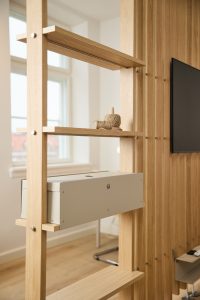 WoodUpp rumdeler i lyst egetræ med lodrette lameller og tre tynde hylder; midterhylden bærer en diskret, grå WoodUpp kabelkasse til ledningsstyring, og øverste hylde har garnnøgle. Stilrent, skandinavisk design der adskiller stuen fra arbejdsområdet, med synlige metalbeslag, rund ventilationsåbning og naturligt lysindfald.