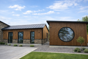 Billedet viser to moderne bygninger med flotte WoodUpp facadeløsninger. Den højre bygning er fuld beklædt med lodrette, bruntonede trælameller og har et stort rundt vindue. Den venstre bygning kombinerer de samme WoodUpp træpaneler på den øvre del med en rustik stenbase under en blå himmel.