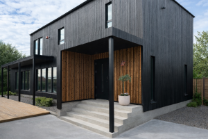 Billedet viser et moderne hus med sort træfacade. Det overdækkede indgangsparti er fremhævet med WoodUpp beklædning. Disse WoodUpp paneler har lodrette lameller i en varm, naturlig egetræsfarve med tydelige, rustikke knaster, hvilket skaber en flot kontrast til den sorte hoveddør og betontrappen nedenfor.