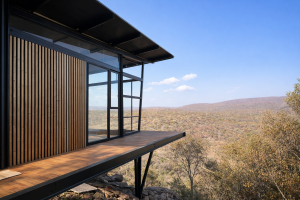 Dette billede viser en moderne arkitektonisk bygning med en udkraget træterrasse. Facaden til venstre er beklædt med smukke, lodrette WoodUpp trælameller i en varm nuance, der står i kontrast til de sorte metalrammer og det store glasparti. Udsigten er over et tørt, kuperet landskab under en klar blå himmel.